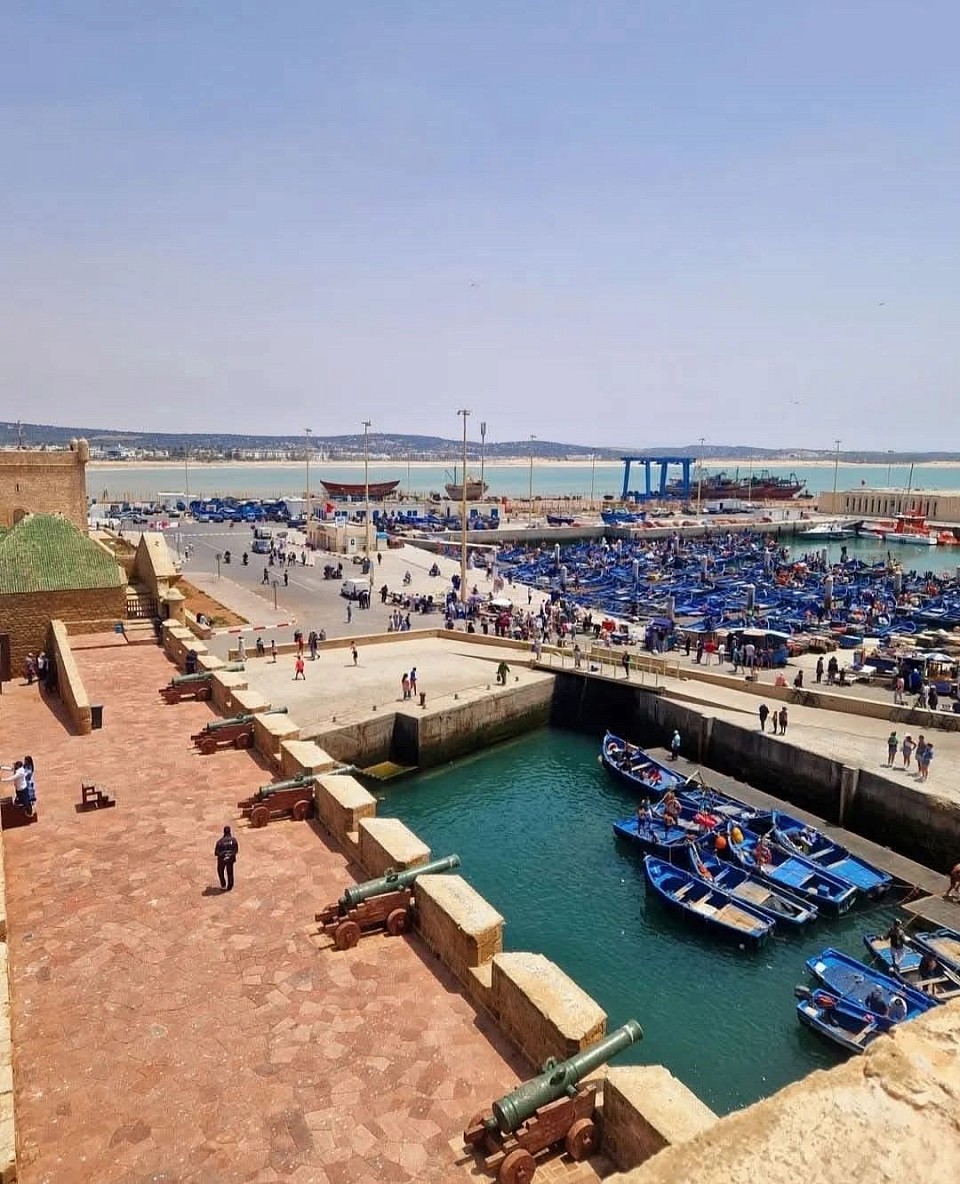 Essaouira Day Trip from Marrakech Essaouira day trip from Marrakech offers a charming escape to the coastal city known for its vibrant culture, captivating history and picturesque scenery. Departing Marrakech in the morning, you’ll embark on a scenic drive through the Moroccan countryside towards the Atlantic coast. Upon arrival in Essaouira, you’ll be greeted by the charming blue and white ramparts of the city’s medina, which create a striking contrast to the blue sea. As you venture into the medina’s narrow, winding streets, you’ll find yourself immersed in a lively atmosphere filled with colorful shops, bustling markets and the aroma of freshly cooked seafood. Stroll along the ancient ramparts surrounding the city, which offer panoramic views of the ocean and the picturesque harbor filled with traditional blue fishing boats. The refreshing sea breeze and the sound of seagulls overhead create a serene atmosphere as you explore the charming streets lined with art galleries, craft shops and quaint cafes.  Essaouira beach is a popular place for relaxation and water activities. Feel the sand between your toes as you take a leisurely stroll along the shoreline or enjoy a refreshing dip in the Atlantic waters. You may even get the chance to witness local fishermen skillfully maneuvering their boats or practicing the ancient art of net fishing. Seafood lovers will delight in the abundance of fresh seafood available in Essaouira. Savor a delicious lunch at a local restaurant, where you can indulge in the city’s famous grilled fish and other local specialties. Afterward, take your time to continue exploring the city’s cultural highlights, such as the Skala de la Ville, a fortification offering stunning views of the coastline, or the lively Place Moulay Hassan, where you can soak up the lively atmosphere and observe the local way of life. At the end of the day, you’ll embark on the journey back to Marrakech, taking with you memories of Essaouira’s charm, coastal beauty, and vibrant cultural scene.  PICK UP: Marrakech Hotel/Riad DROP OFF: Marrakech Hotel/Riad INCLUSIONS: Pick up and drop off at Marrakech Riad Transportation by air-conditioned minibus Professional driver Day trip to Essaouira EXCLUSIONS: Food and drinks